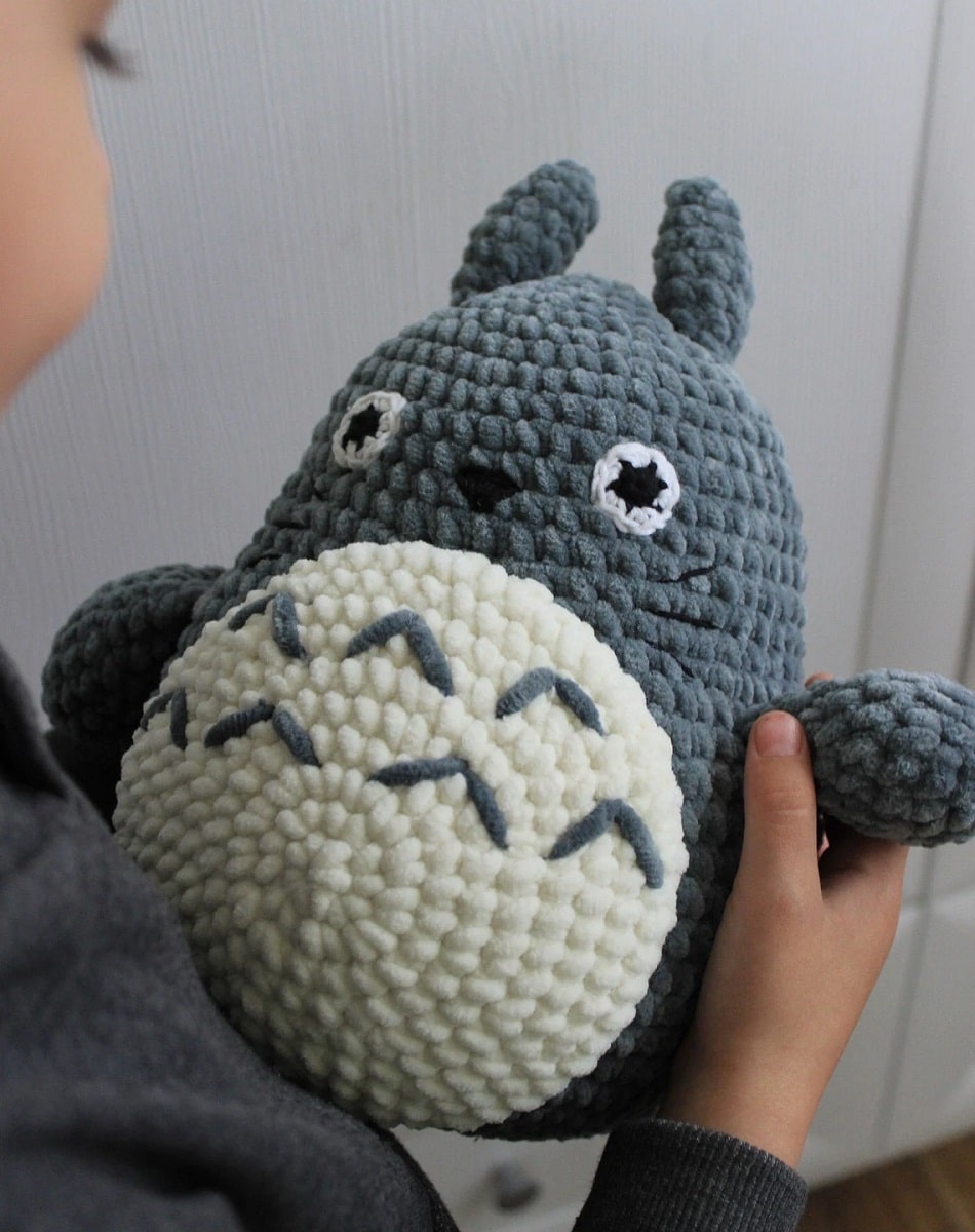 Large stuffed crochet Totoro toy with a textured dark flue face and body and white belly with small blue stitches on the top.