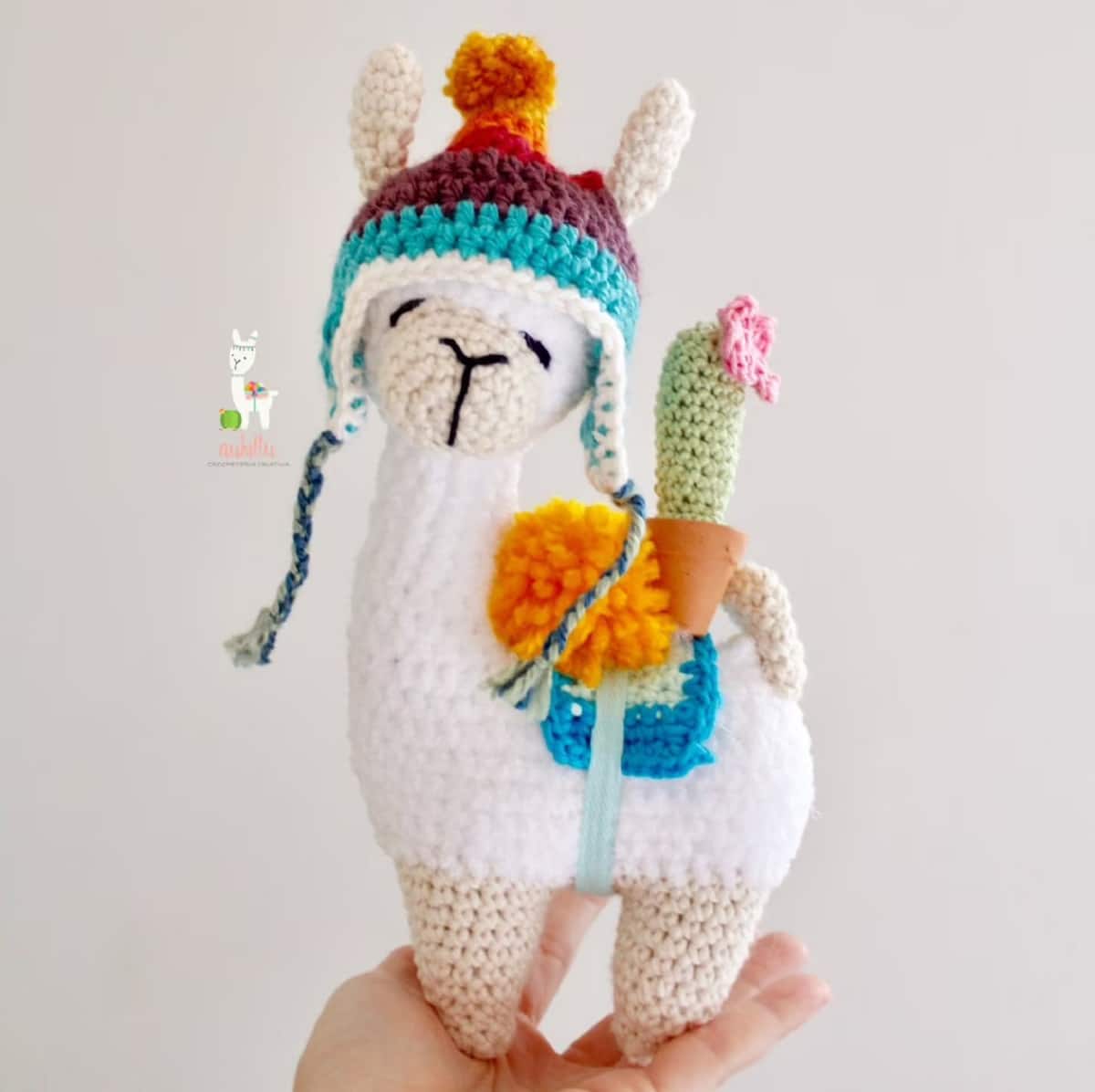 White crochet llama wearing a blue and brown hat with braids either side, a blue seat around its middle with a cactus and orange flowers standing on its back.