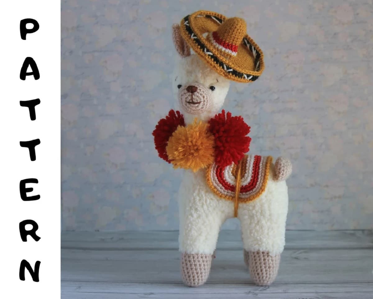 White crochet fluffy llama wearing a brown sombrero, with brown and orange fluffy bobbles around its neck and a multi-colored seat on its back.