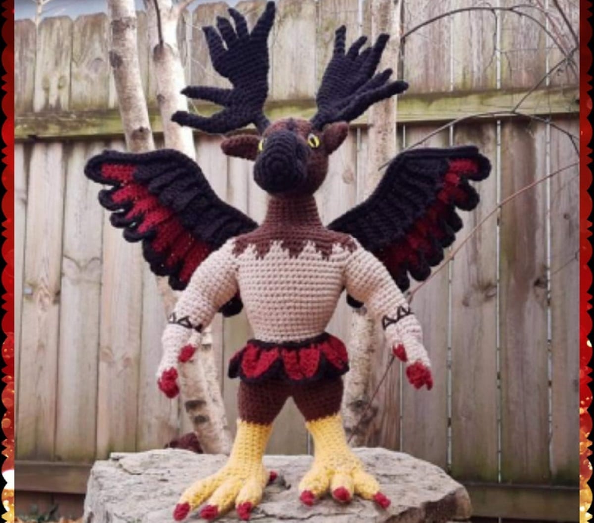 Crochet Pamela, a mountain spirit with antlers, wings, and animal feet standing on a rock in front of a wooden fence. 