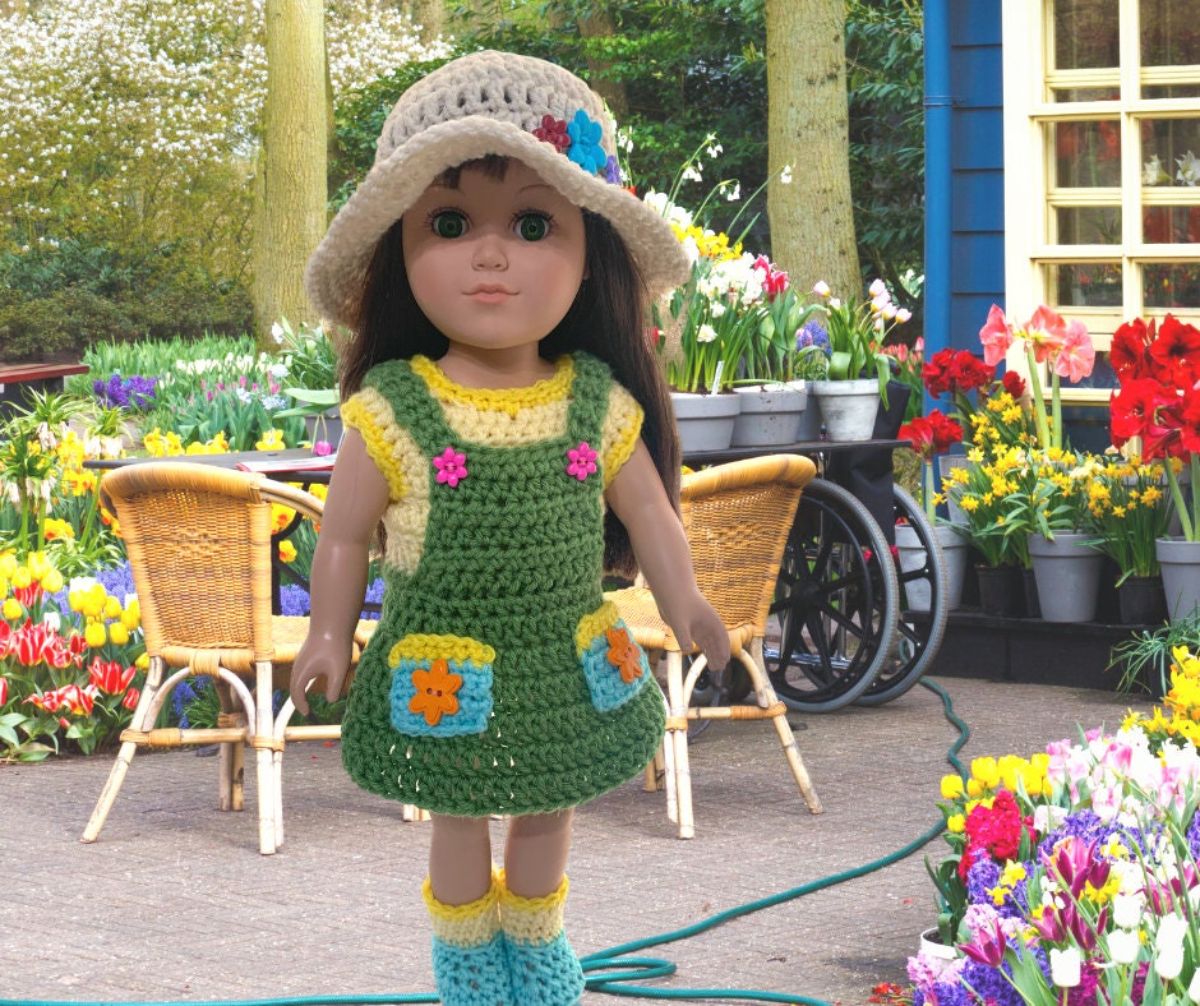 Brunette American girl doll wearing a crochet sun hat with a yellow t-shirt and green dress over the top with little pockets.