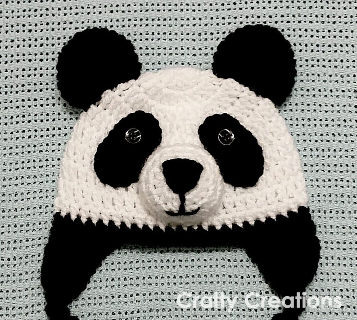 Black and white crochet panda beanie with black flaps and string to fasten on a pale green crochet blanket.
