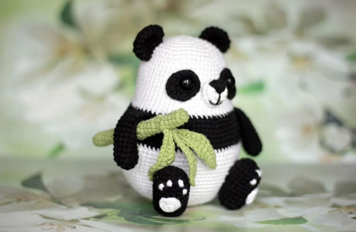 Small black and white crochet panda sitting down with a crochet green bamboo shoot tucked under his arm. 