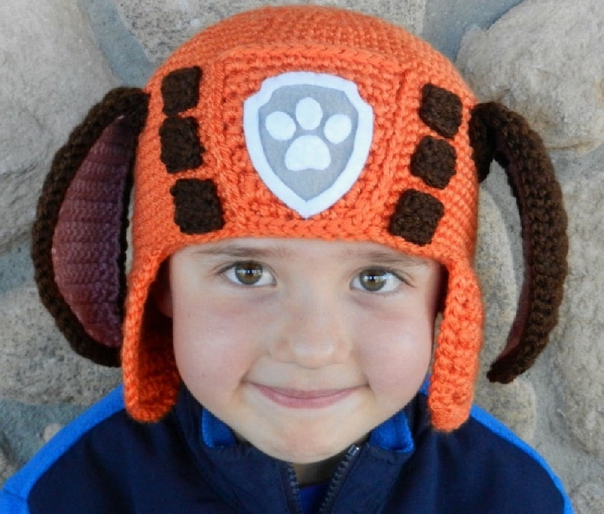 Young boy wearing an orange crochet hat with orange earflaps, large brown ears either side, and a gray Paw Patrol logo in the center of the hat.
