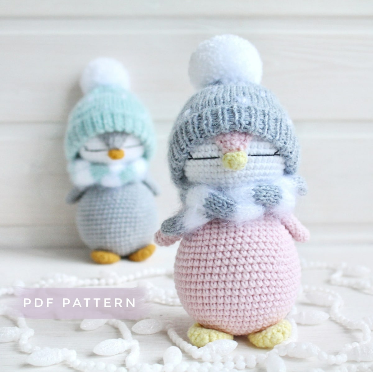 Crochet penguin with a pale pink body and white face wearing a gray bobble hat and gray and white scarf with a smaller penguin in the distance wearing a blue hat.