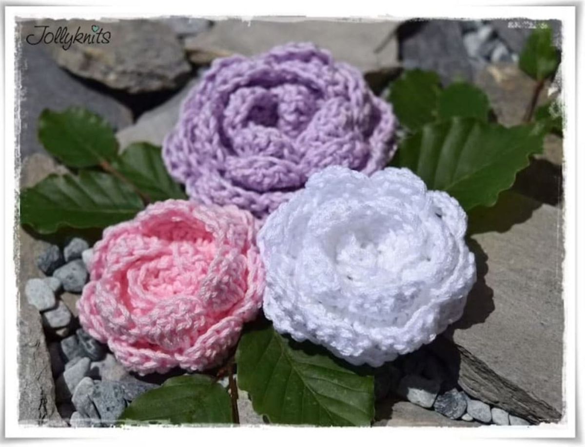 Crochet peonies in white, purple, and pink sitting on top of artificial leaves and small gray stones with a gray background.