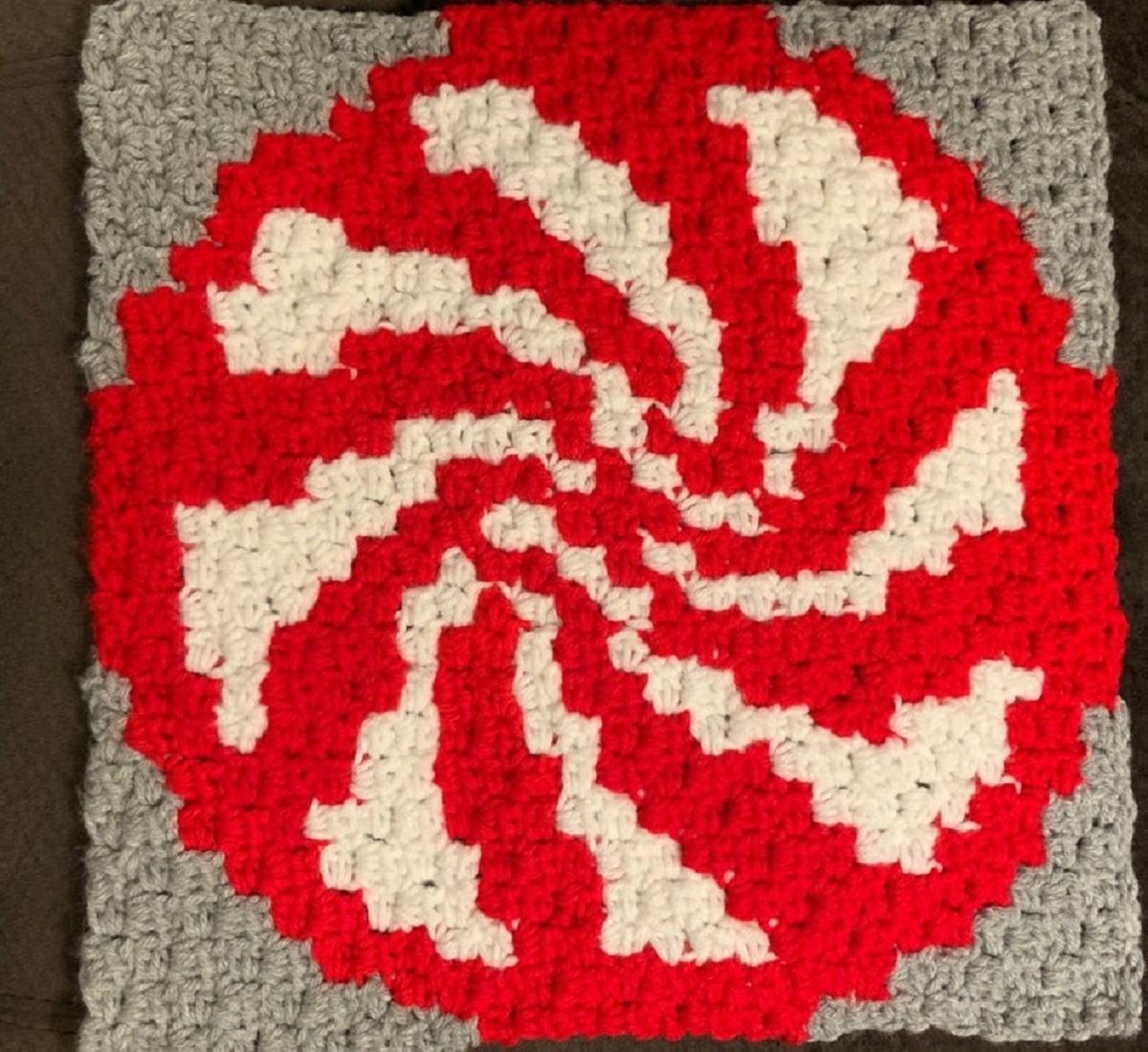 A gray crochet cushion cover with a large red circle with red swirls in the center on a dark wooden background.