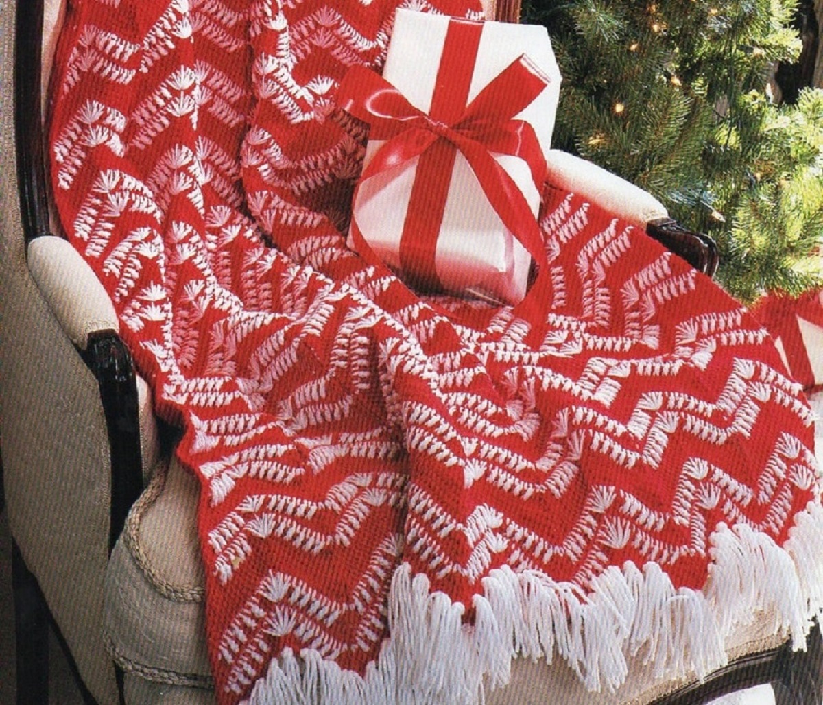Large red crochet Afghan with white zig zag stripes across the blanket and white tassels on the bottom draped over a white chair.
