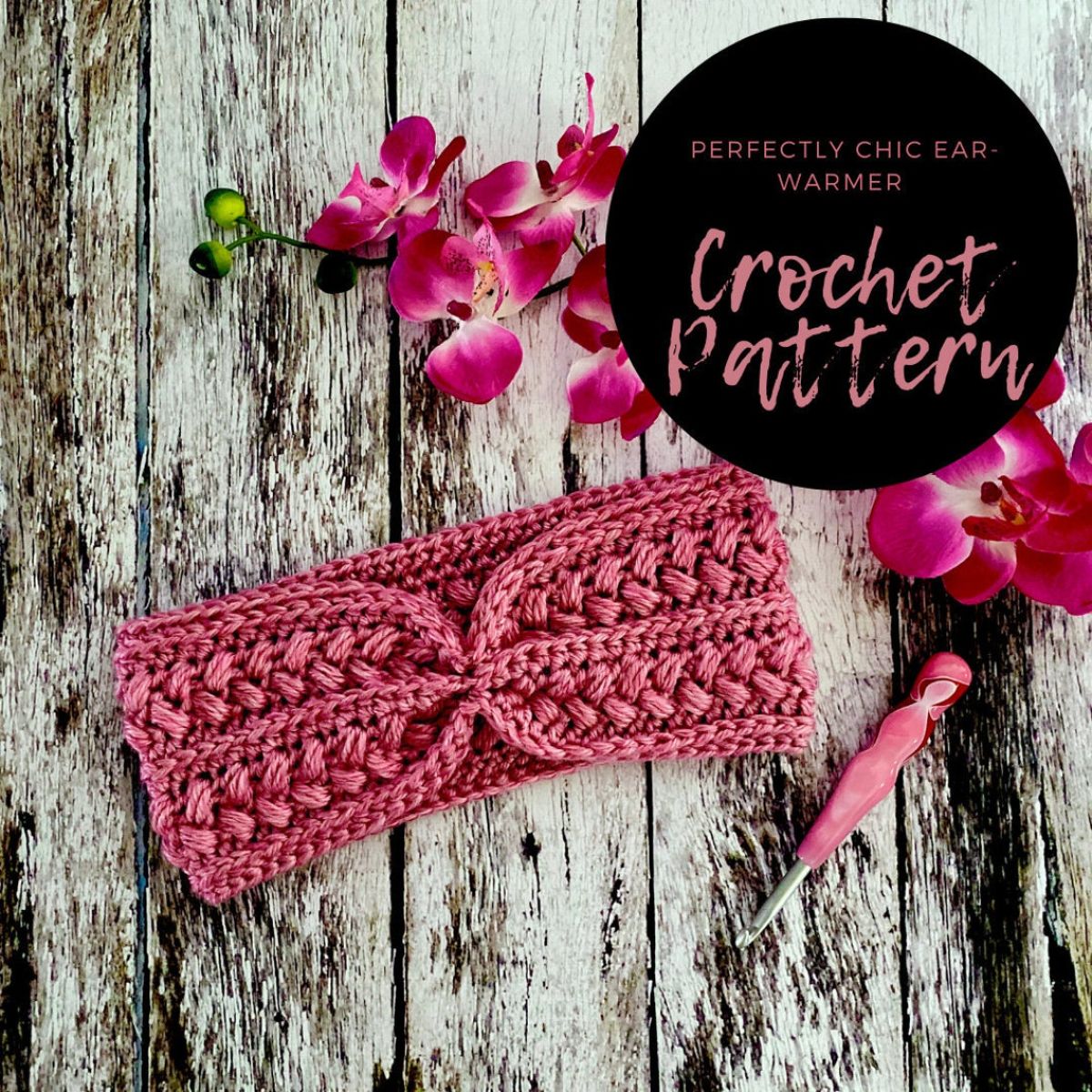 Pink crochet ear warmer with small bobble design that meets in the middle on a wooden background with a pink crochet hook and orchid next to it.