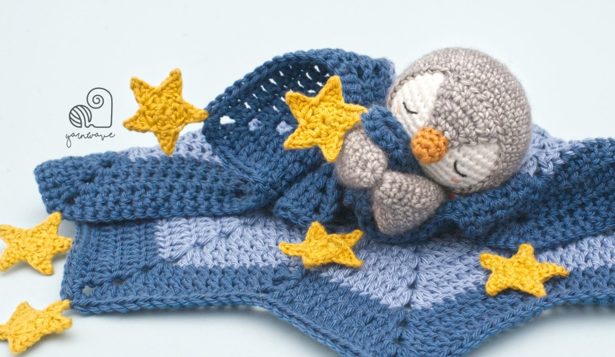 Gray penguin with an orange nose and its eyes closed tucked under a blue star shaped crochet blanket with gold stars scattered around them.