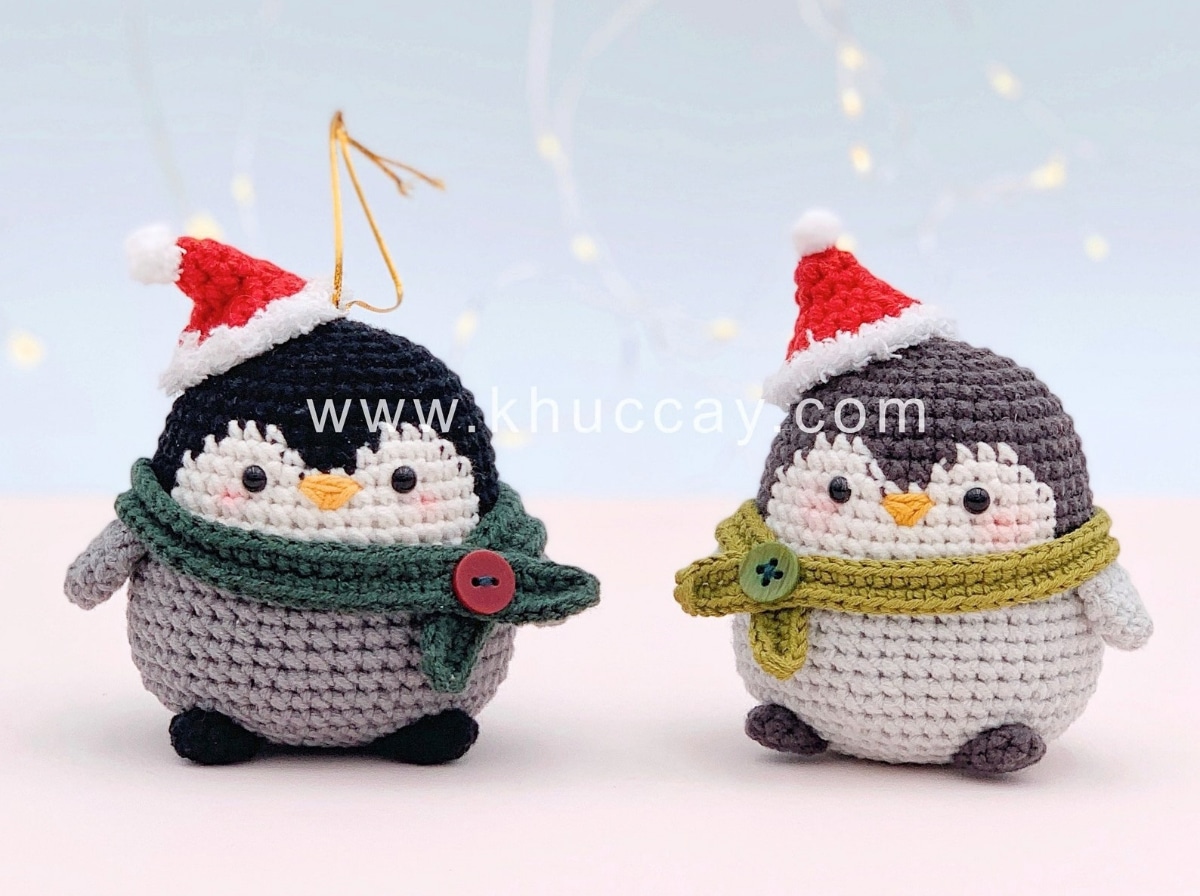 A black and white baby penguin wearing a red Christmas hat and green scarf next to a gray baby penguin wearing a red Christmas hat and light green scarf.