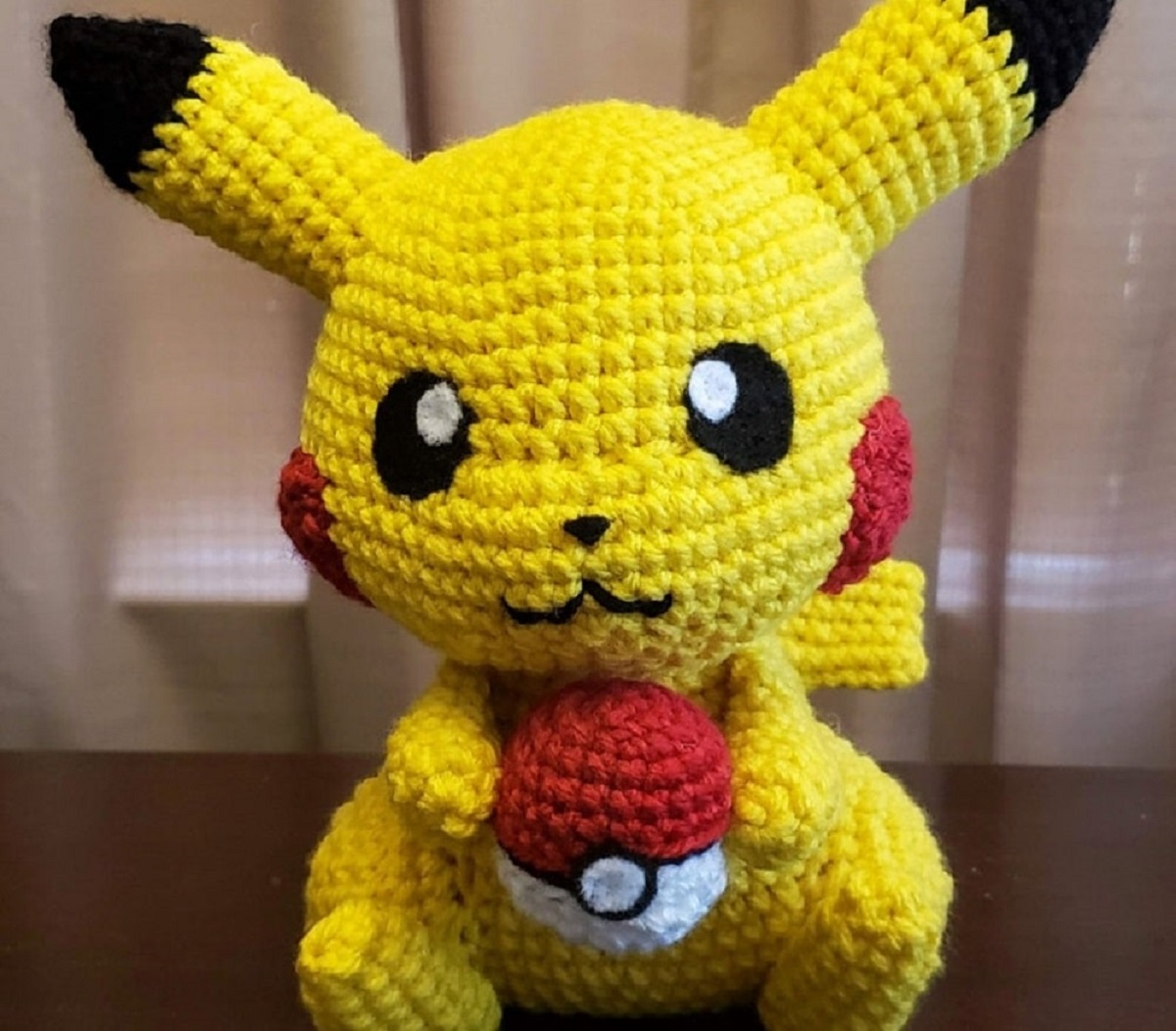 Large stuffed yellow crochet Pikachu toy holding a red, white, and black Pokeball sitting on a dark wooden floor.