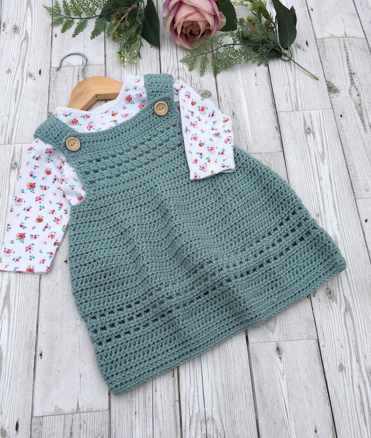 Green crochet pinafore baby dress with a button on either strap with a white floral top underneath.