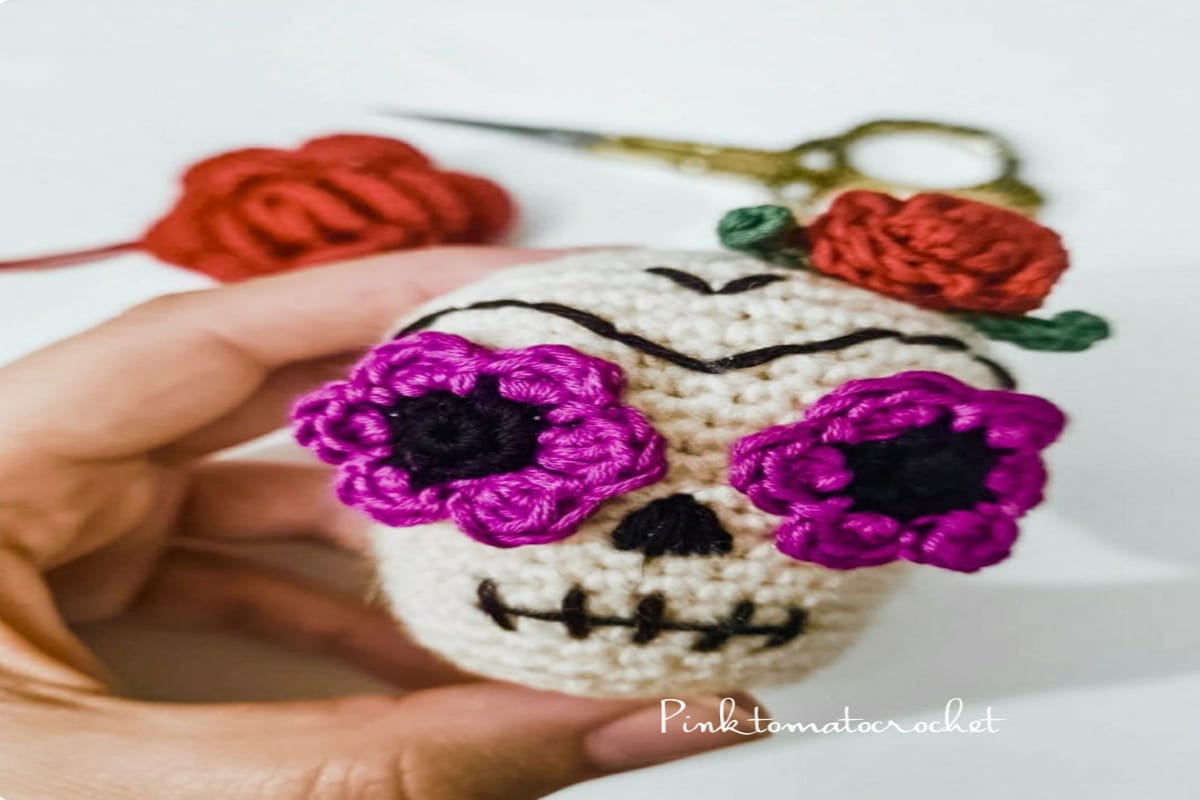  A hand holding a small white skull with pink flowers for eyes and red crochet roses in the background.