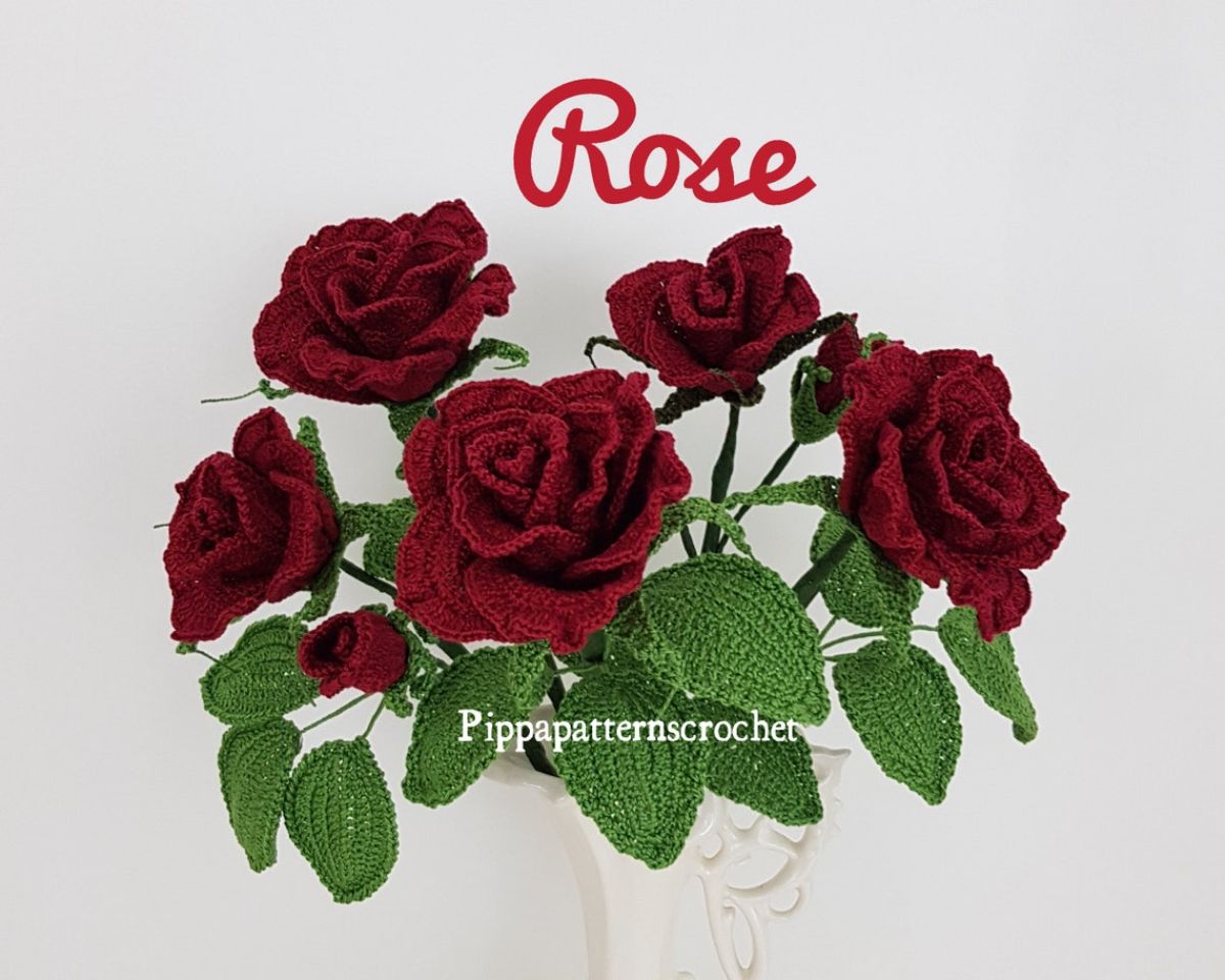 A vase of dark red crochet roses with green leaves hanging over the side on a white background.