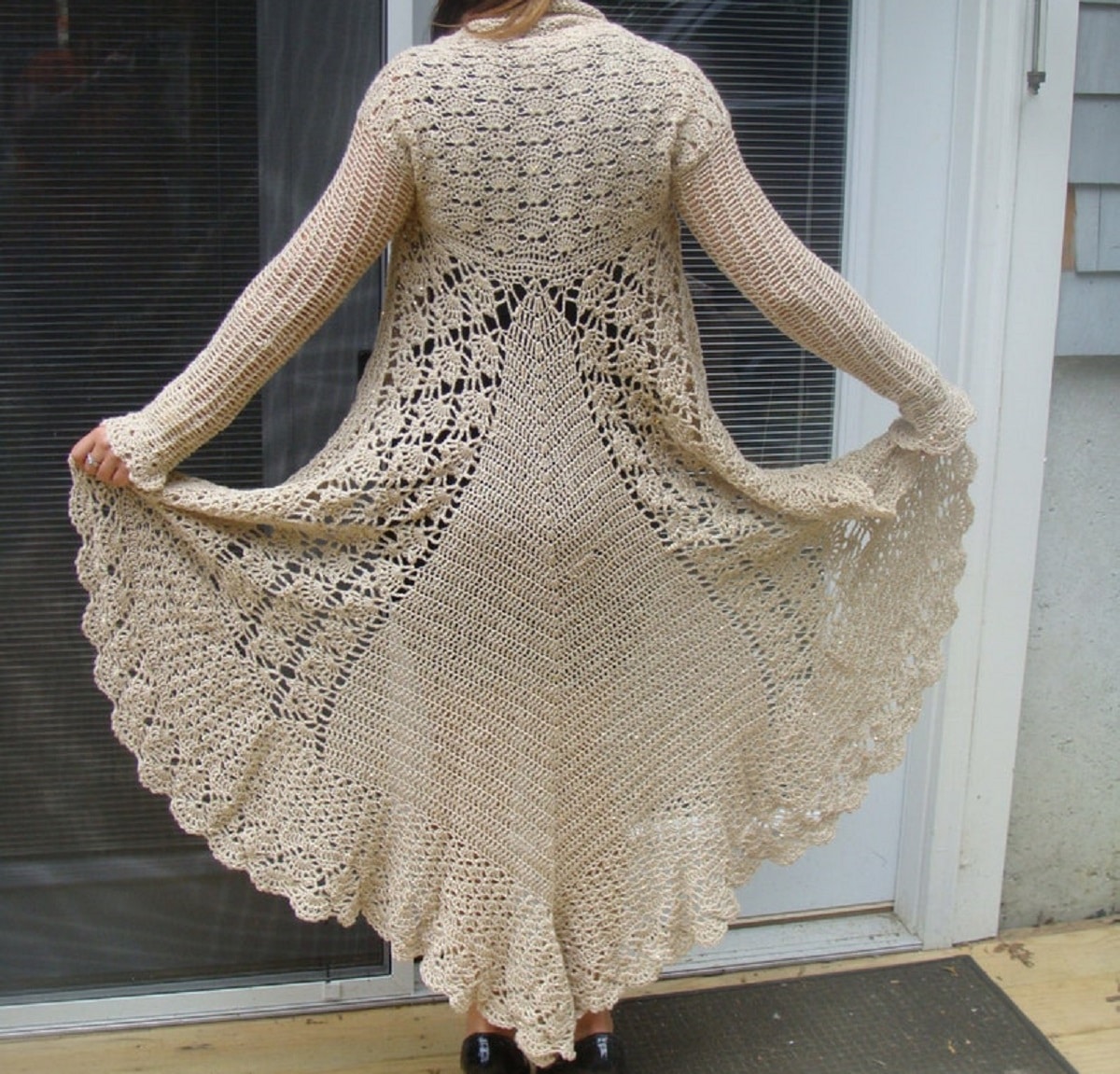 The back of a woman wearing a beige long sleeved full length crochet sweater standing in front of a glass door.