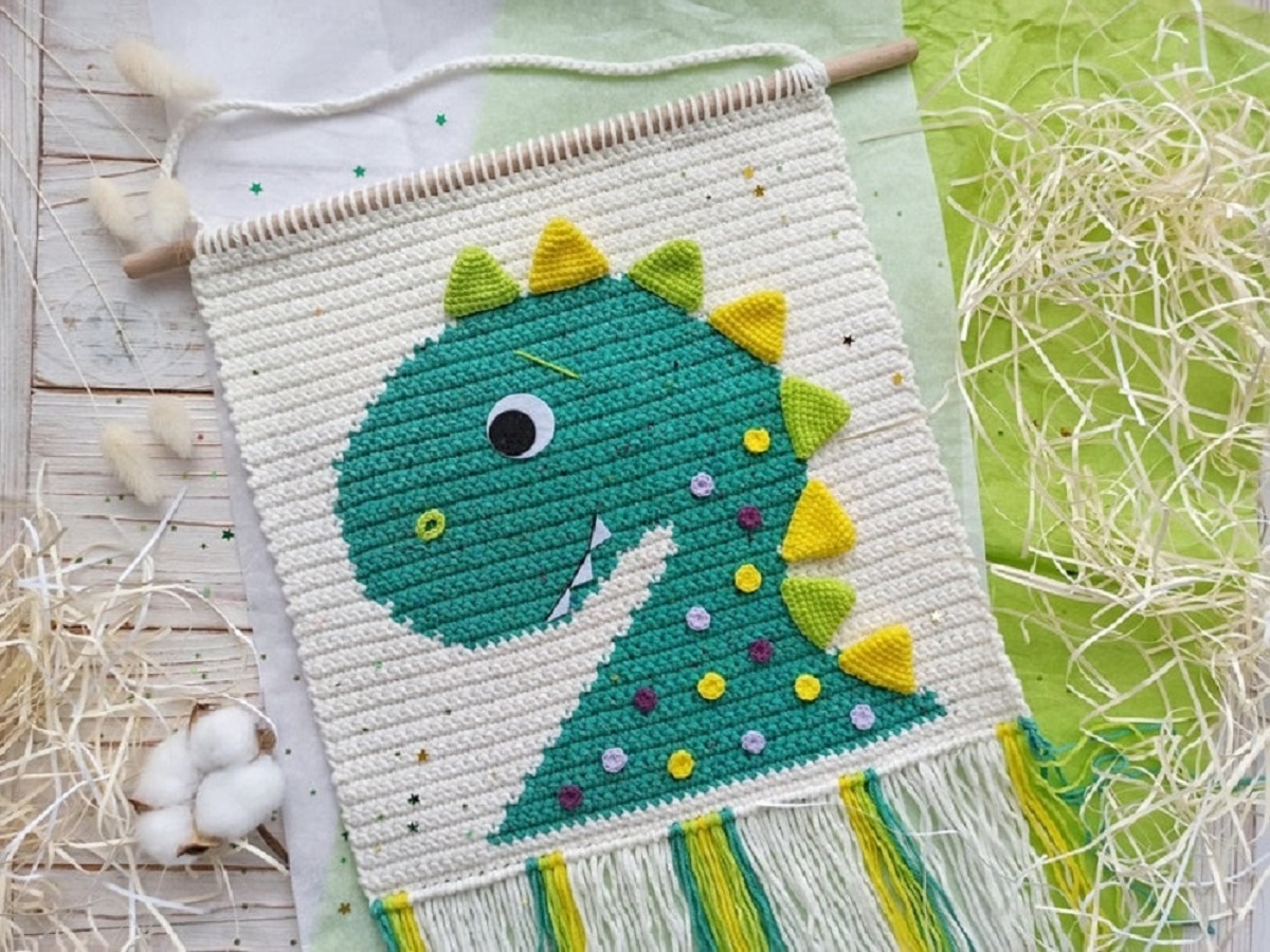 A white crochet wall hanging with a green dinosaur with yellow and green spikes down its back and purple, yellow, and white spots on its neck.