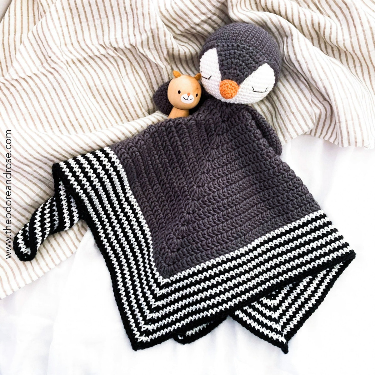 Black crochet blanket with white diagonal stripes on the bottom third with a penguin head stitched at the top laying on a bed with a cat cuddly toy.