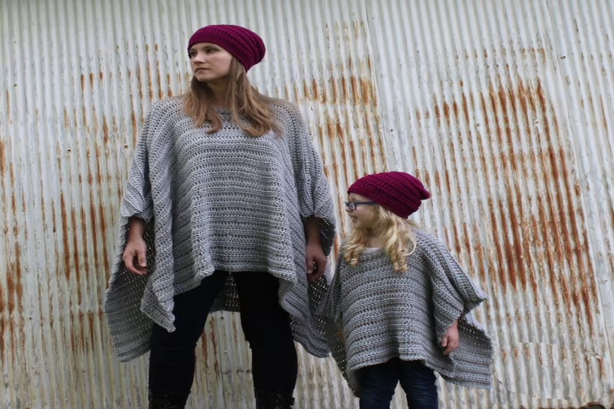 A mother and daughter in matching gray crochet ponchos and red beanie hats against a rust colored background.