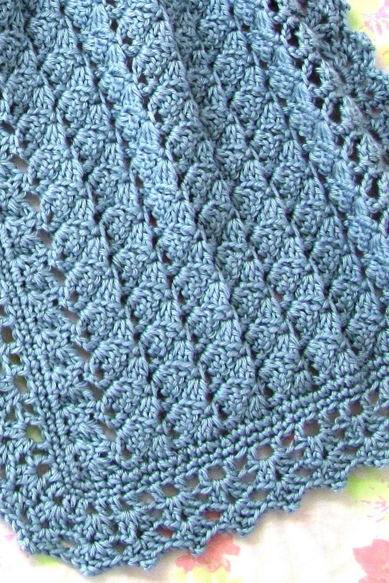 A blue crochet prayer shawl with a lace trim on all sides on a light colored background.