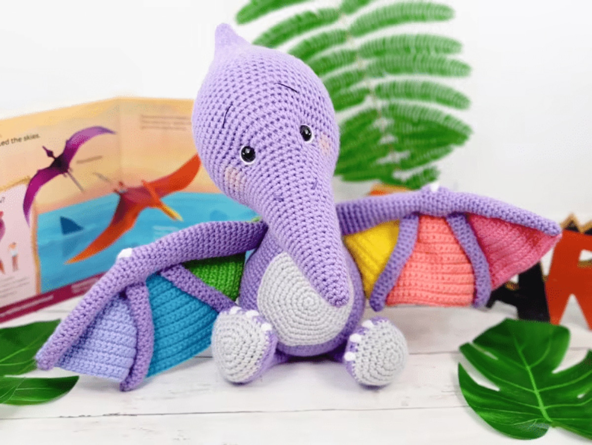 Purple crochet Pterodactyl with a white stomach and feet, and pink, orange, yellow, green, blue, and purple triangles on its wings.