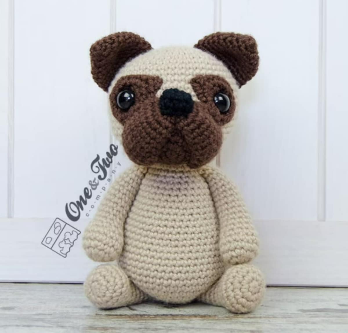 Crochet stuffed Pug with a beige body and brown eyes, nose, and years sitting up against a white wooden door.