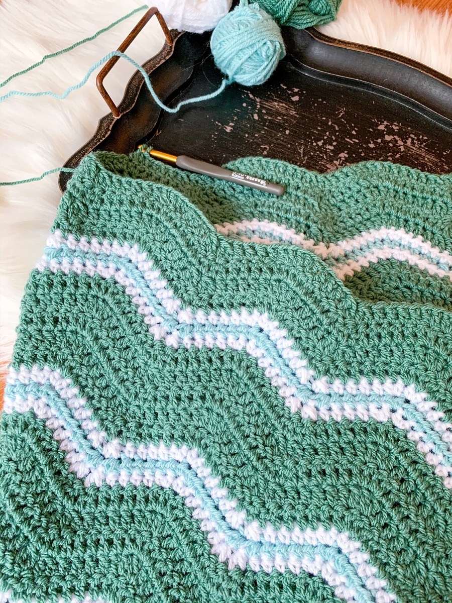 Green, blue, and white crochet wave blanket with horizontal waves on a metal tray with blue yarn and crochet hooks next to it. 