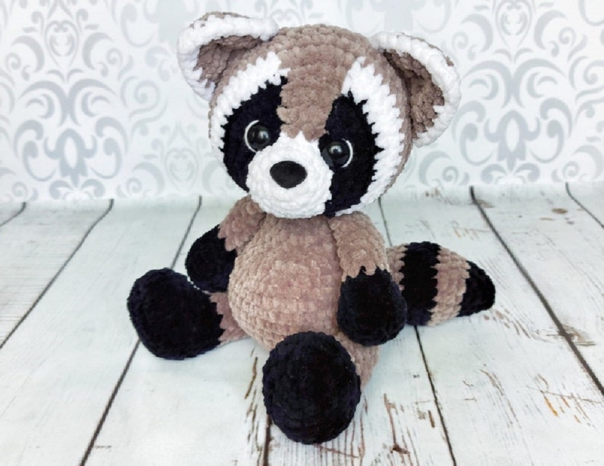 Small crochet stuffed raccoon with a light brown body, dark brown and white eyes, dark brown feet, and a striped tail.
