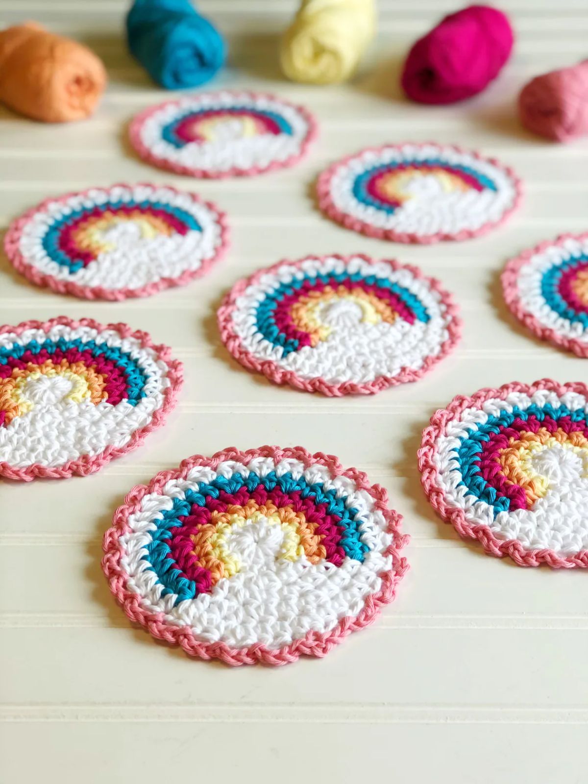 Small white crochet coasters with a pink trim and a rainbow in the top half spread out on a pale wooden table.