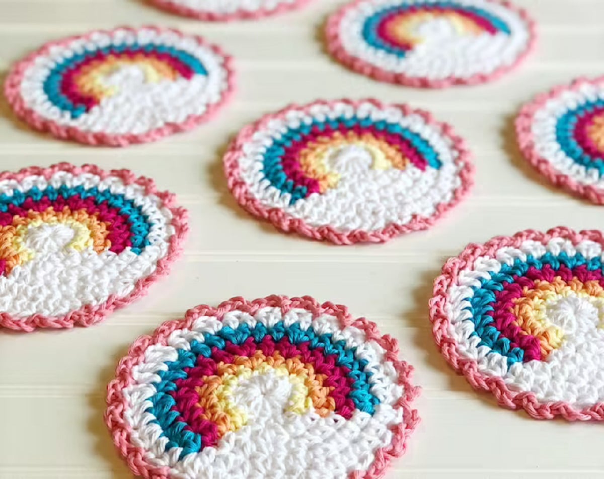 Several white crochet coasters with a rainbow arched across the top half spread across a pale wooden background.
