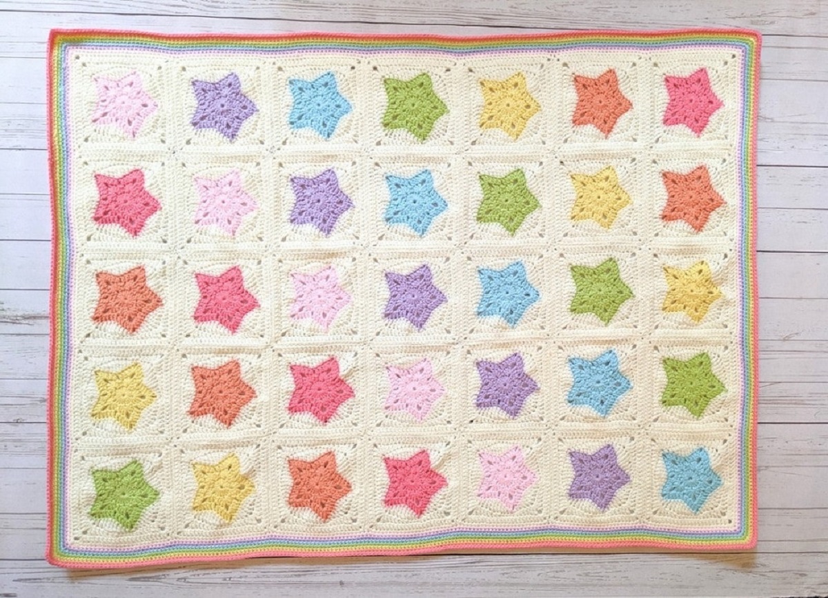 Large cream crochet blanket with rainbow colored stars stitched into squares and a red and green trim around all sides of the blanket.