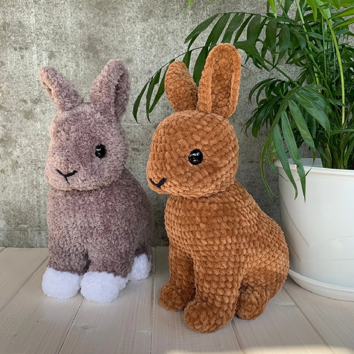 Two large life-like brown crochet stuffed bunnies sat next to each other on a white wooden floor next to a plant pot.