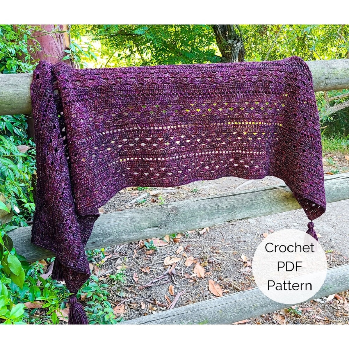  Dark purple rectangular crochet shawl draped over a wooden fence next to a muddy path and some leaves.