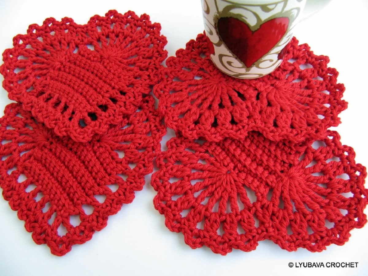 Four crochet red heart coasters stacked on top of each other with a white cup with gold detailing and a red heart on top. 