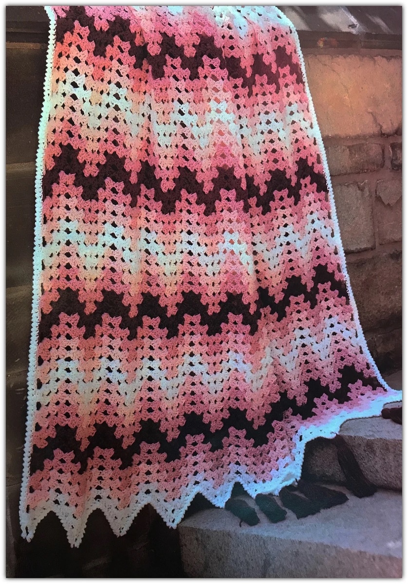 Large black, orange, pink, and cream crochet Afghan with zig-zag horizontal stripes draped over a brick wall. 