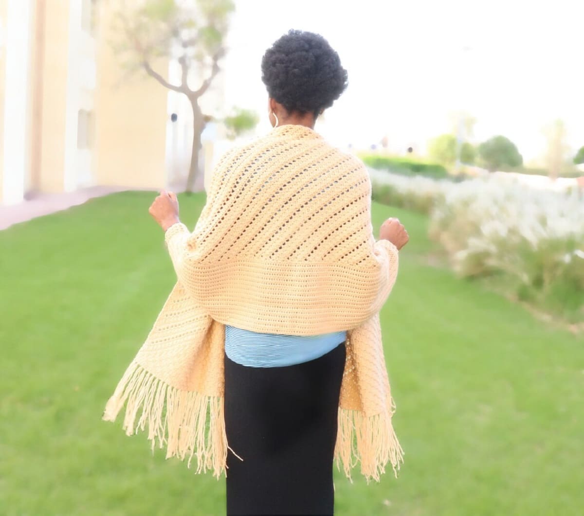 The back of a woman wearing a yellow crochet prayer shawl with yellow tassels along the bottom.