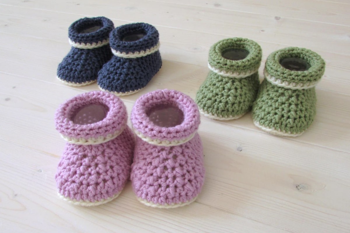 Pink crochet booties with a roll top and cream soles in front of sage green and black booties in the same style.