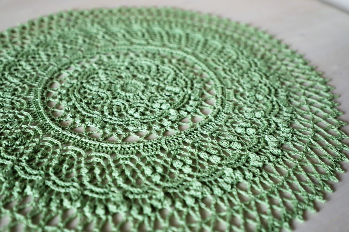Round green crochet rug or decoration with a lace like pattern spreading outwards on a soft white background.