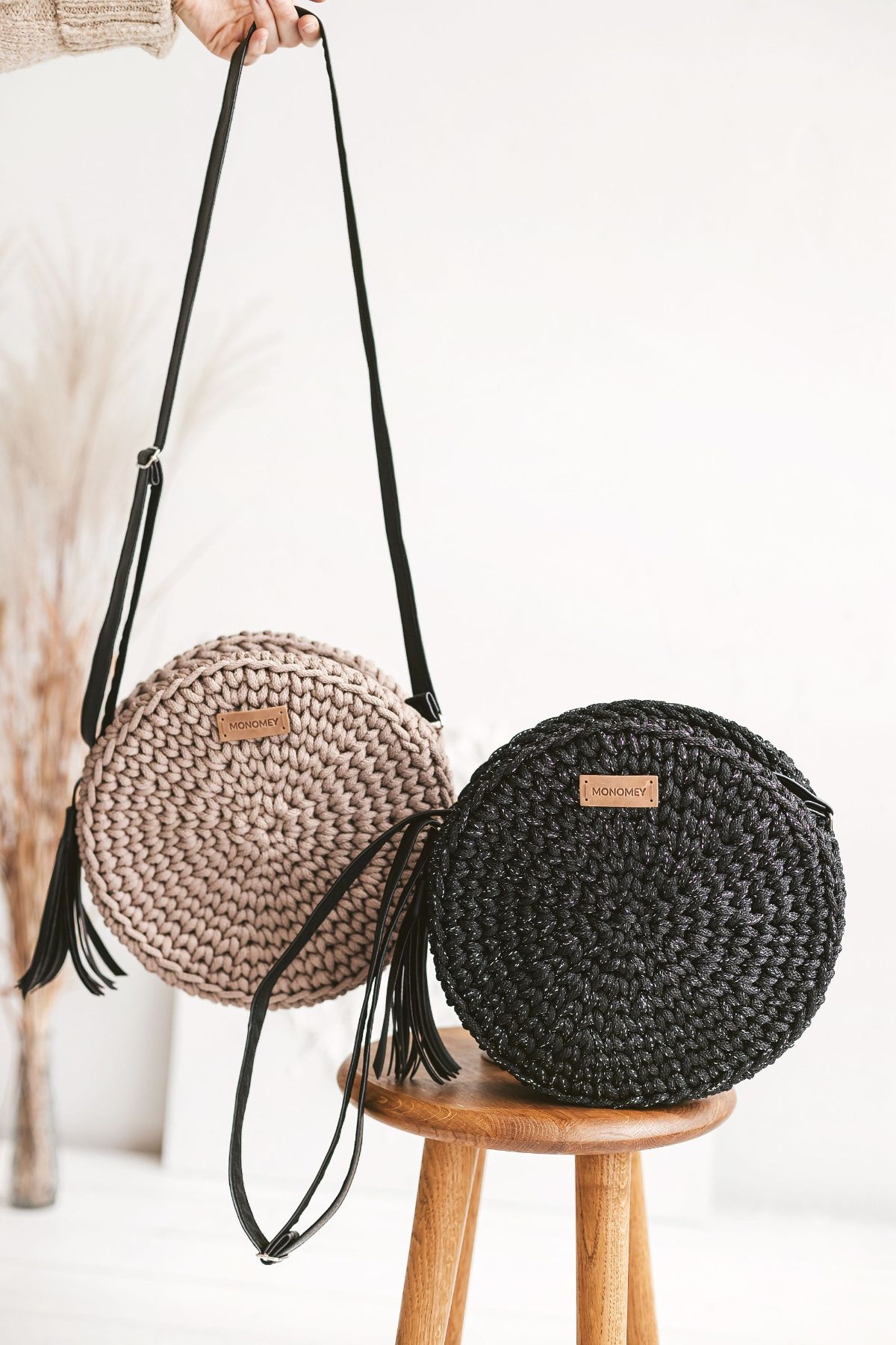 A black round crochet bag with a long strap and tassle on a wooden stool with the same bag in pale pink hanging behind it.