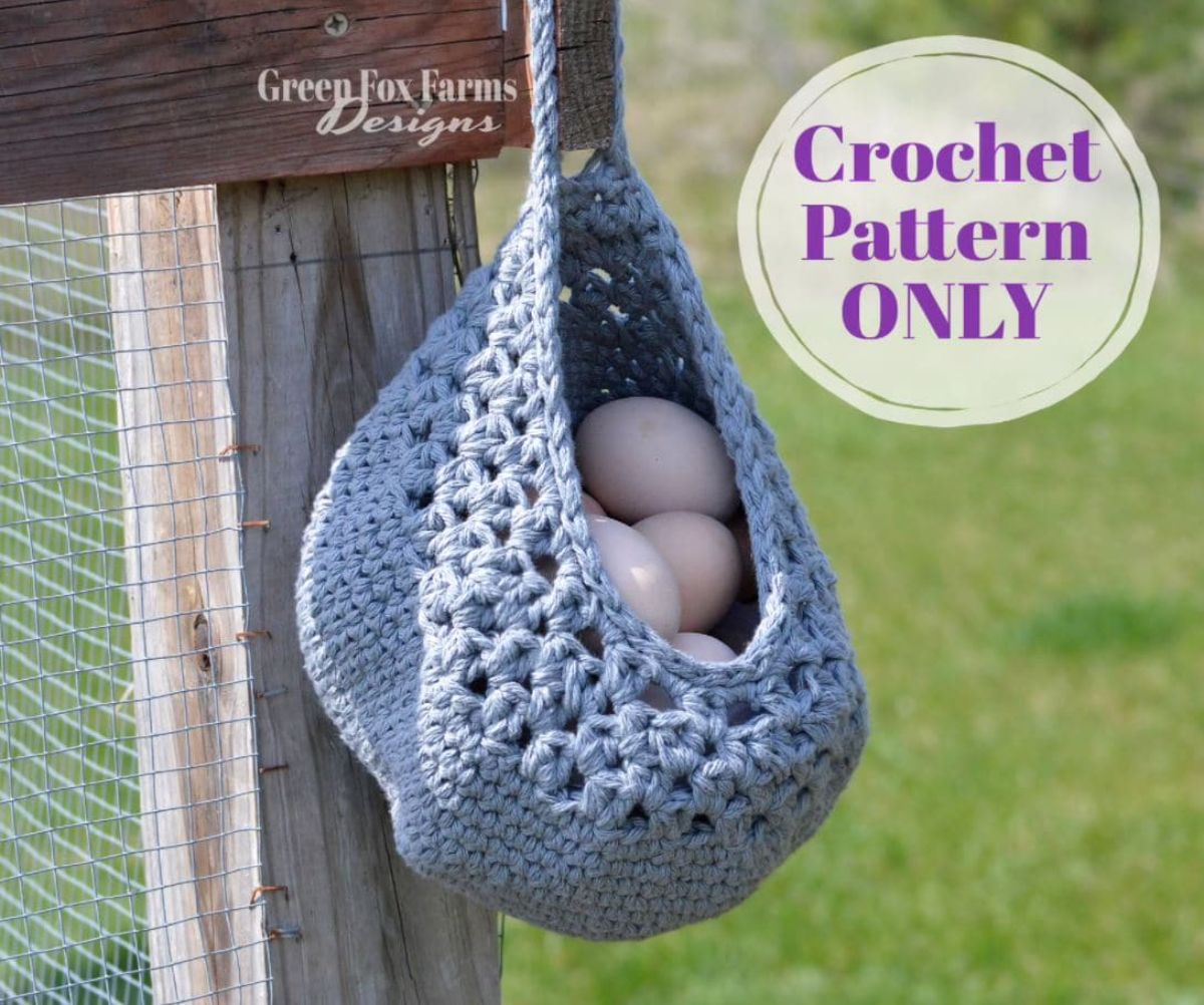 Pale blue crochet basket filled with chicken eggs hanging from a wooden and mesh fence with grass behind. 