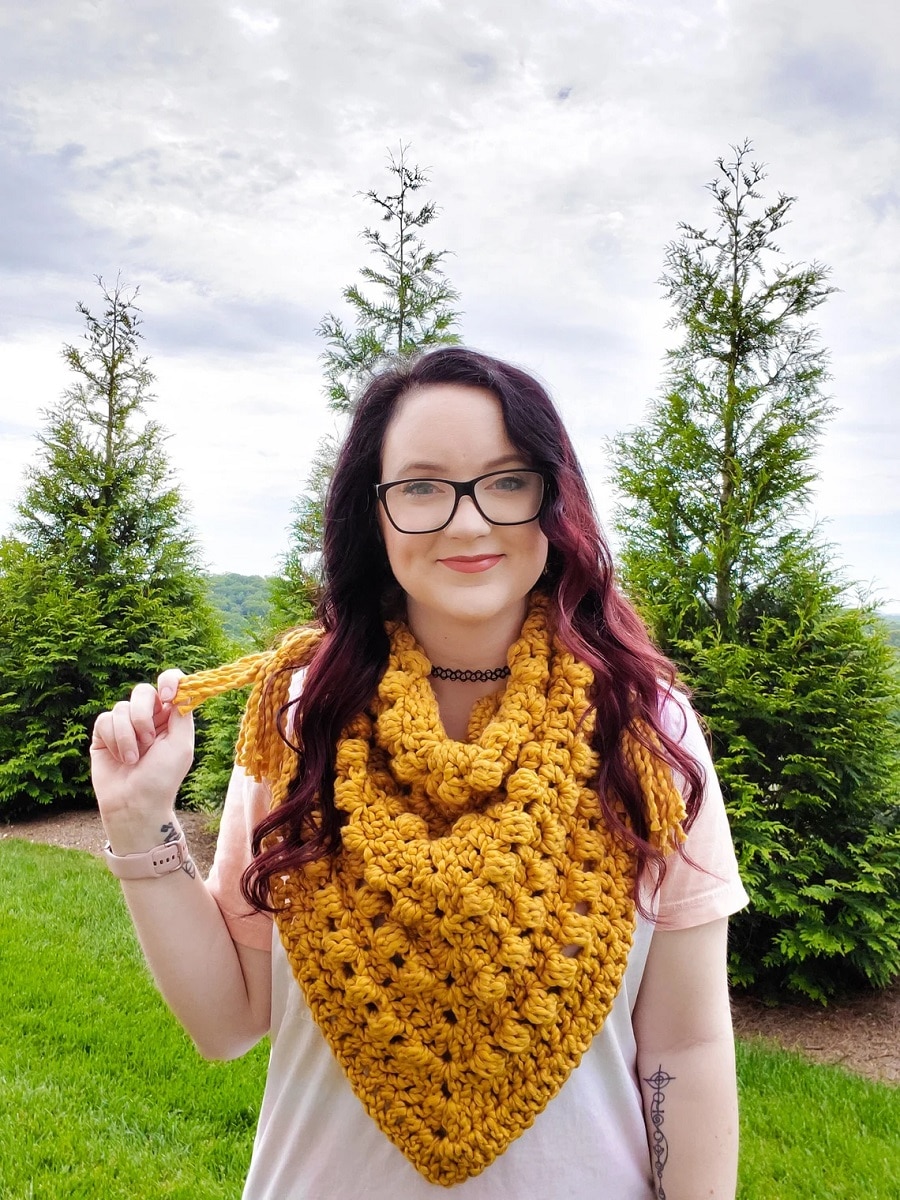 Dark haired woman wearing a yellow crochet triangle scarf with bobbles around the neck and a yellow tassel on the left side.