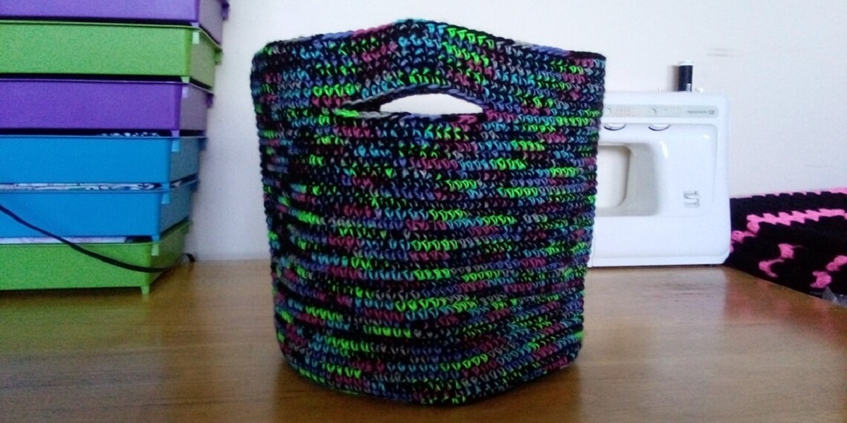 A dark purple, green, and black crochet market style bag with thick handles sitting on a wooden desk.
