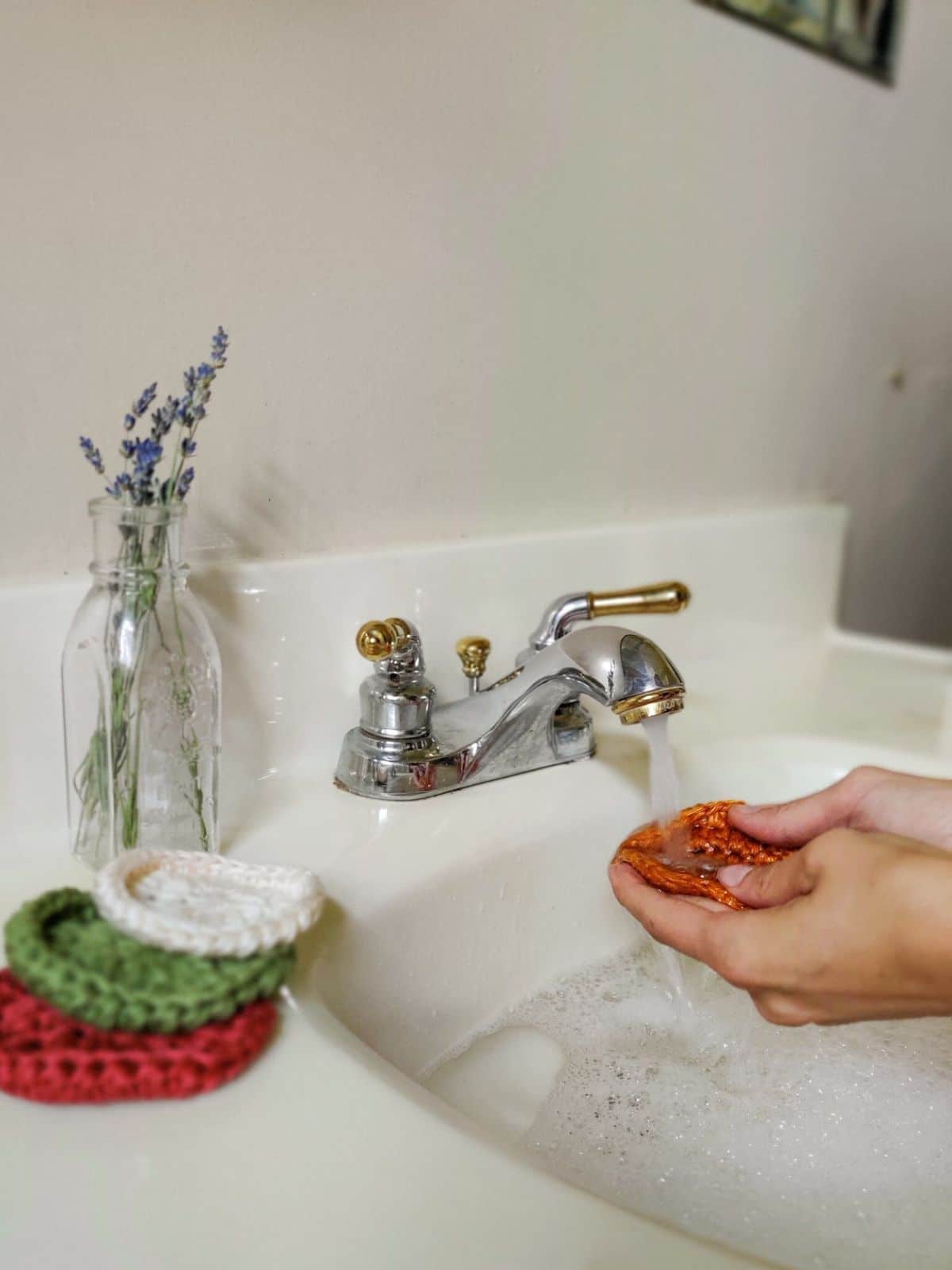 A pair of hands holding a pink crochet scrubbie under a running tap over a sink with a small pile of scrubbies next to it.