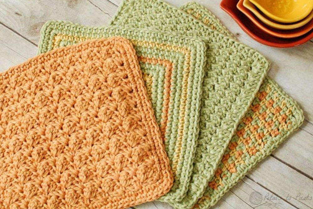 A mustard yellow crochet dishcloth on top of a small pile of green, yellow and orange dishcloths on a wooden table.