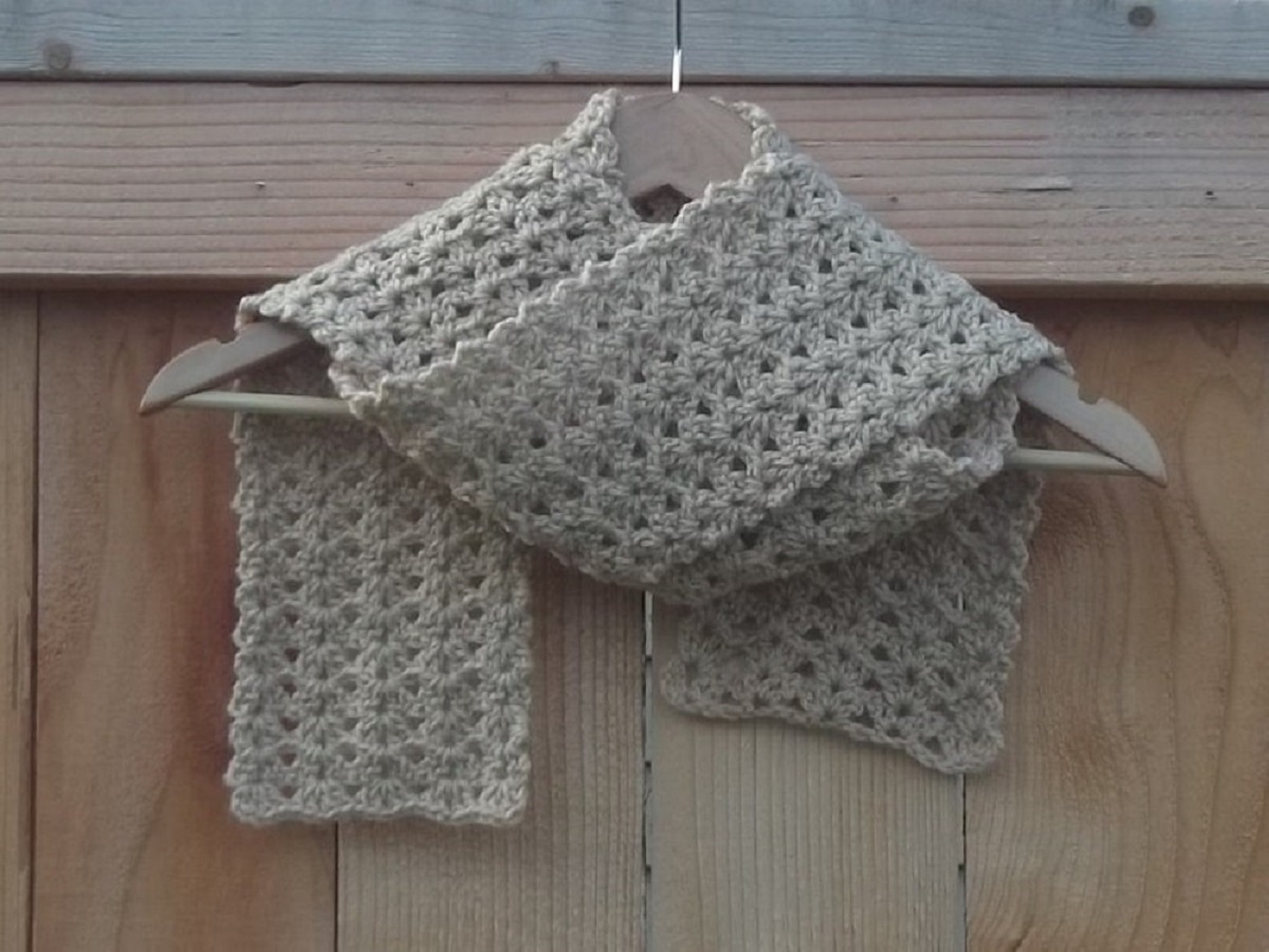A beige colored crochet scarf using a shell pattern folded and draped over a gray hanger hanging from a wooden fence.