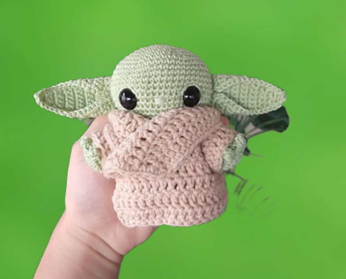 A hand holding a light green crochet baby Yoda in a pale pink robe with an oversized collar on a bright green background.