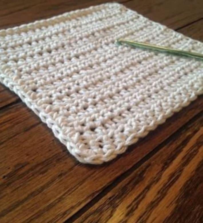 A white crochet dishcloth with a green crochet hook on top of it sitting on a brown wooden table.