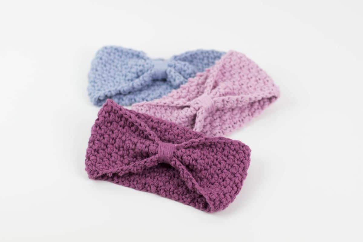 Dark purple, lilac, and pale blue ear warmers with bow design on the front angled together on a white background.