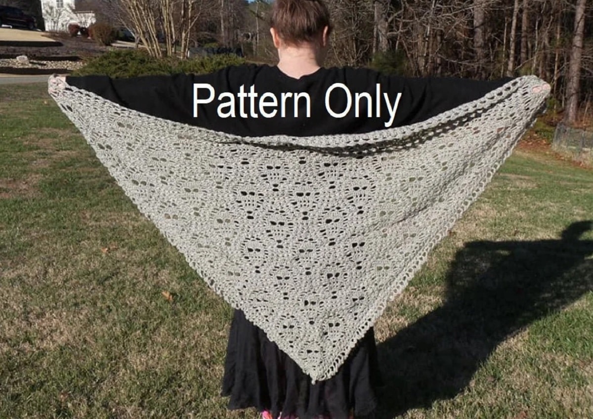 Brunette woman holding a cream crochet diamond shaped shawl with a skull pattern above some short cut grass.