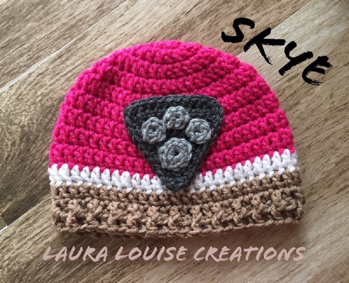 Small crochet pink, white, and brown striped hat with a gray Paw Patrol logo in the center on a wooden background.